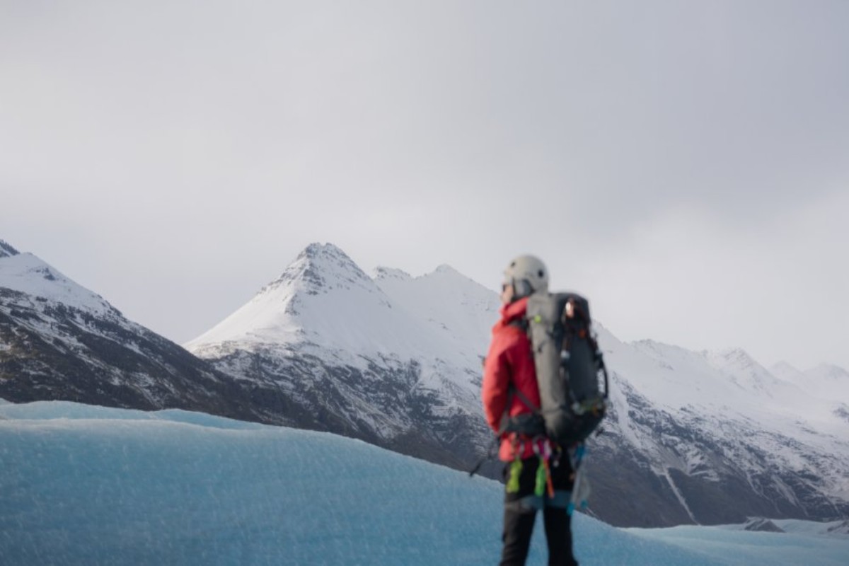 Glacier hiker in a red jacket standing beside blue ice with snowy Icelandic mountains in the background